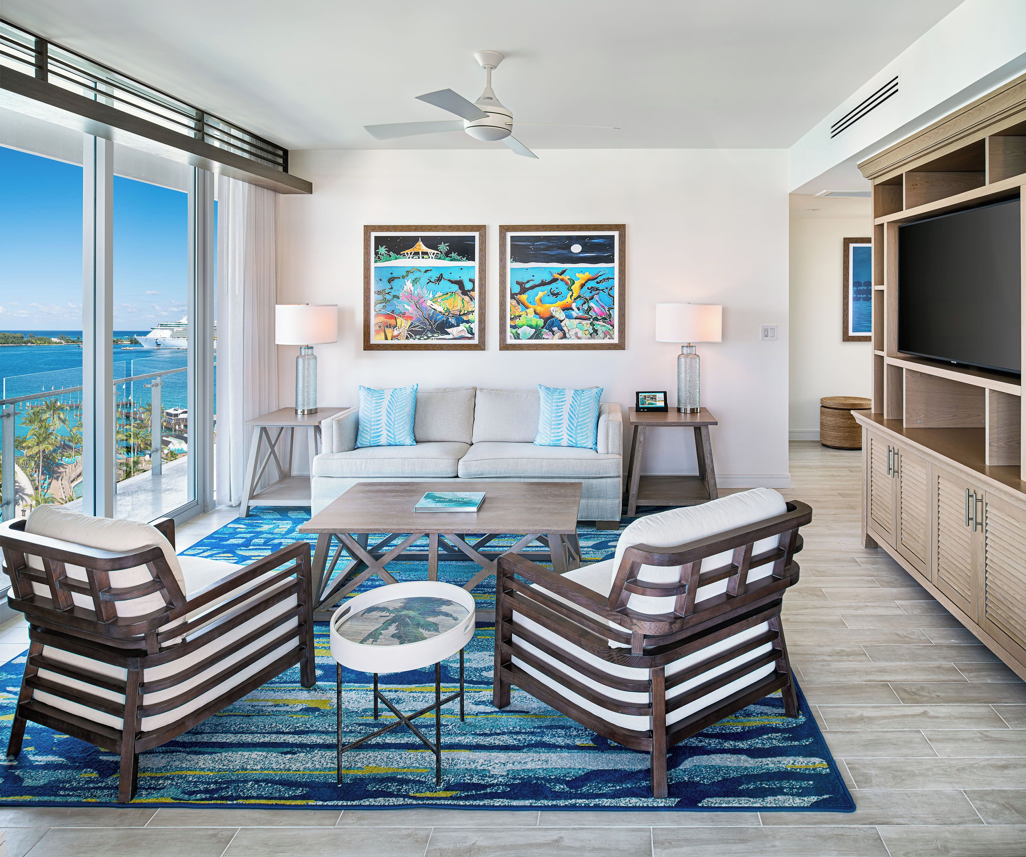 A modern living room with a sofa, two armchairs, a coffee table, and a TV stand. There are two paintings on the wall, a ceiling fan, and a view of the ocean through large windows.