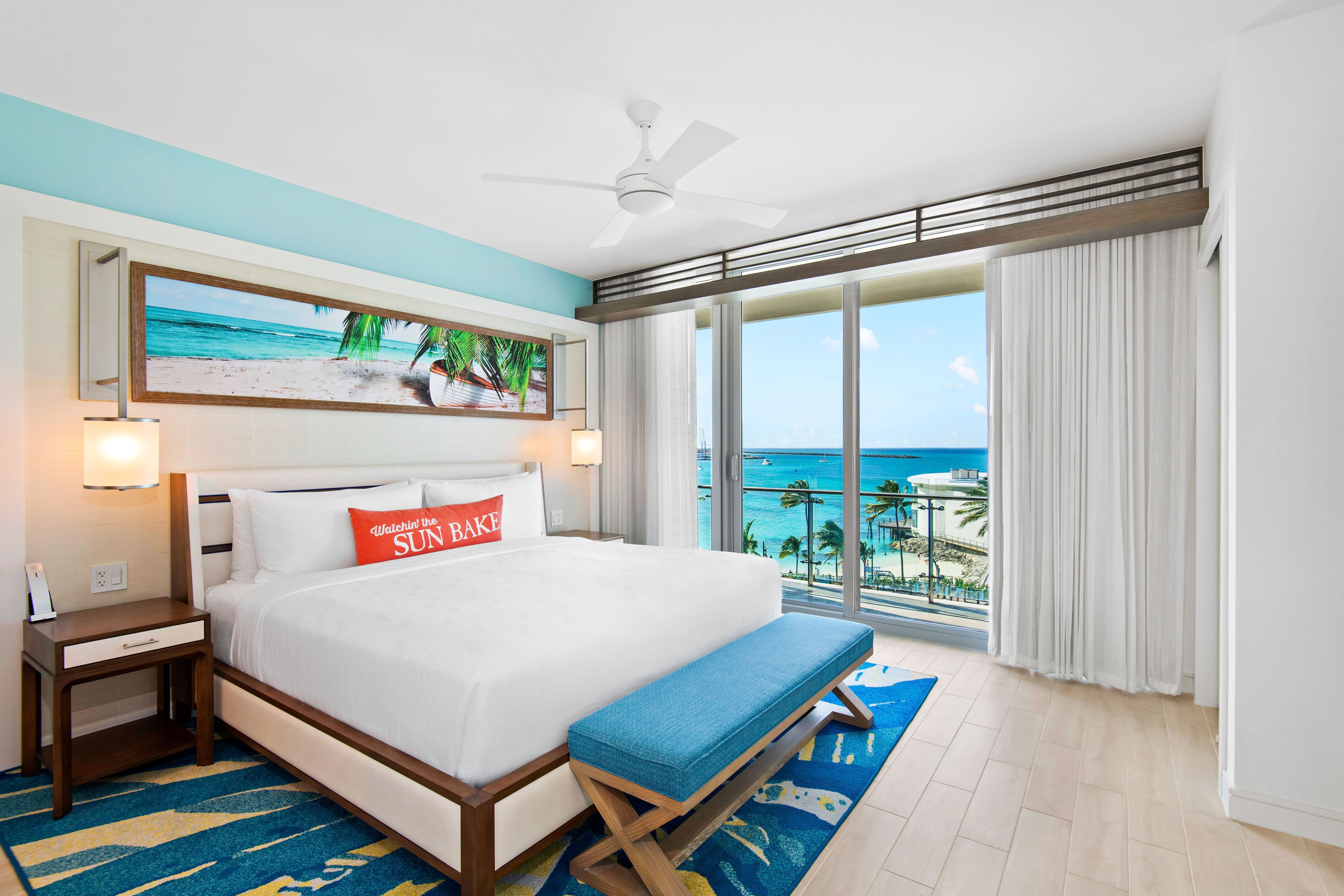 Una habitación de hotel con una cama grande, un banco, un ventilador de techo, un cuadro de playa y vista al océano.