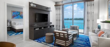 A modern living room with a flat-screen TV, a sofa, a dining area, and a large window with a view of the ocean.