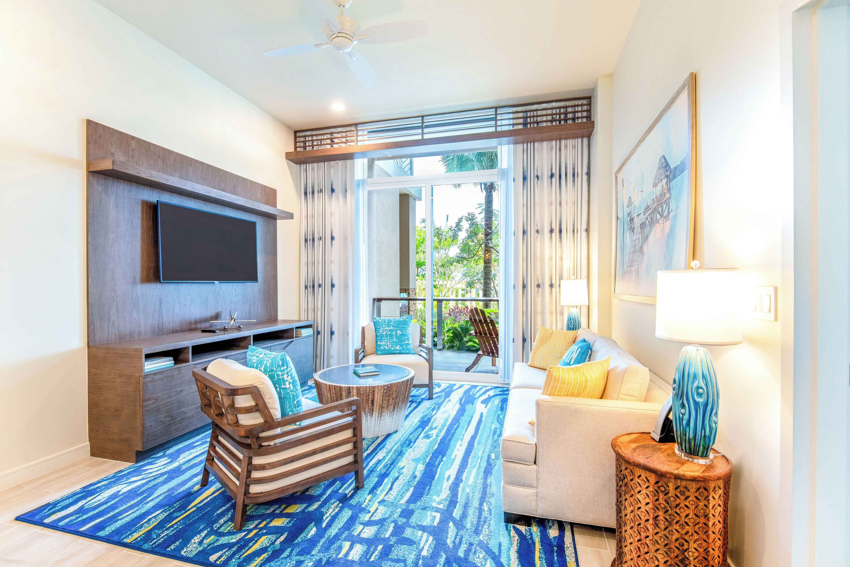 A modern living room with a flat-screen TV, comfortable seating, a coffee table, and a large window with a view of greenery.