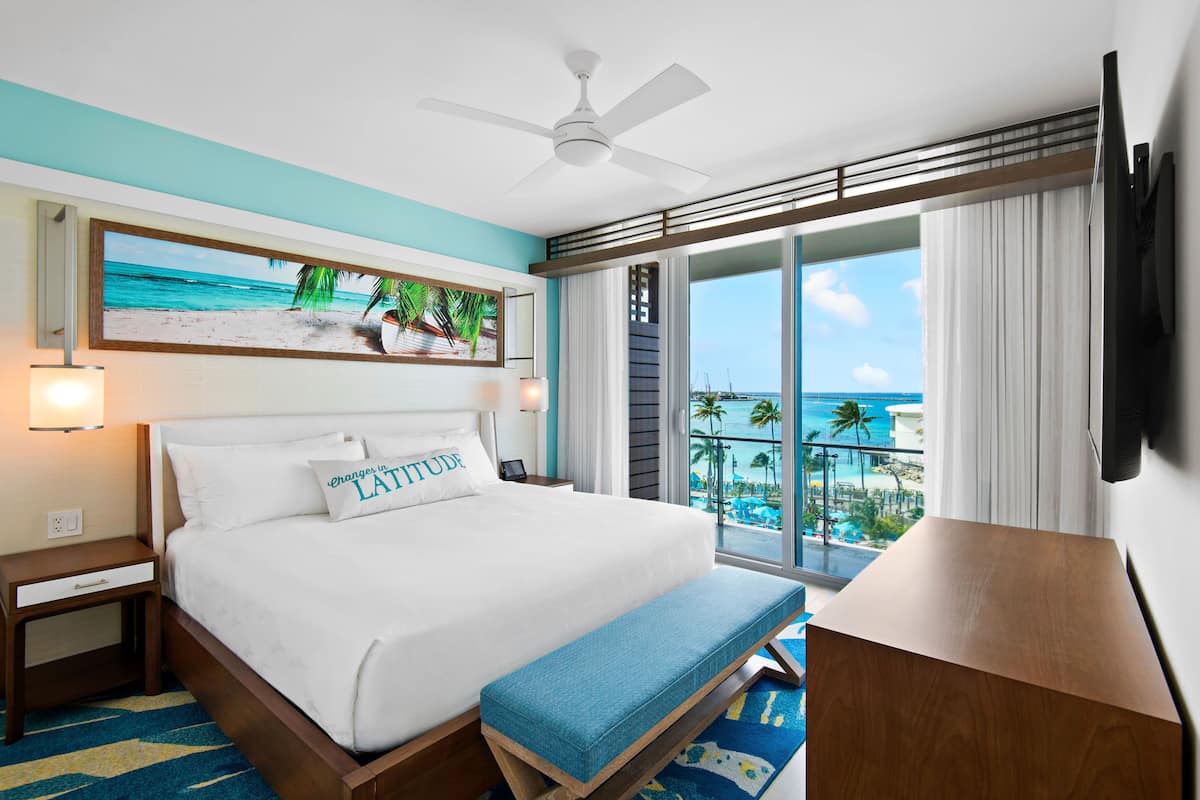 Ein Hotelzimmer mit Bett, einer Bank, einem Deckenventilator, einem Fernseher und einem großen Fenster mit Blick auf das Meer und Palmen.