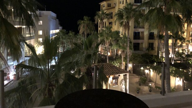 Een nachtzicht van een hotel met meerdere verdiepingen, palmbomen en een klein verlicht gebouw op de voorgrond.
