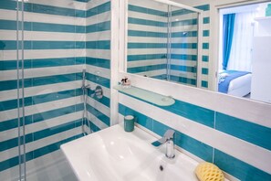 Een badkamer met een witte wastafel, een douchecabine van glas en een spiegel die een bed met blauwe gordijnen weerspiegelt.