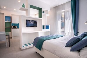 A modern bedroom with a bed, a TV, a desk, and a kitchenette.