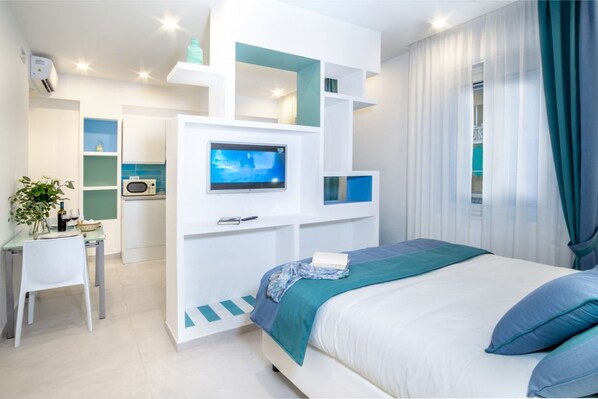 A modern bedroom with a bed, a TV, a desk, and a kitchenette.