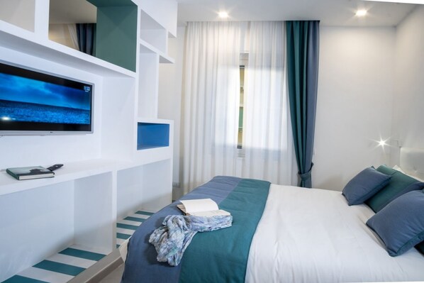 A modern hotel room with a bed, a flat-screen TV mounted on the wall, a window with curtains, and a small shelf with a book and remote control.