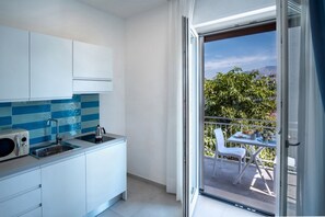 Cocina moderna con armarios blancos, salpicadero de azulejos azules y microondas integrado. Una puerta corrediza de vidrio da a un balcón exterior con una mesa, sillas y vistas a la vegetación y colinas.