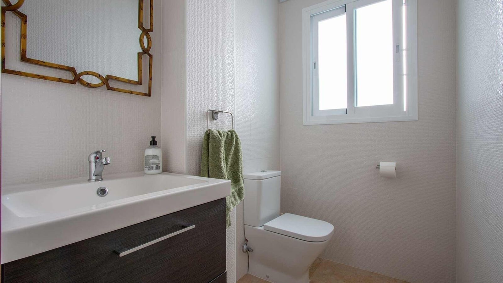 Une salle de bain avec un lavabo blanc, un meuble en bois foncé, une serviette accrochée au mur, des toilettes et une fenêtre.