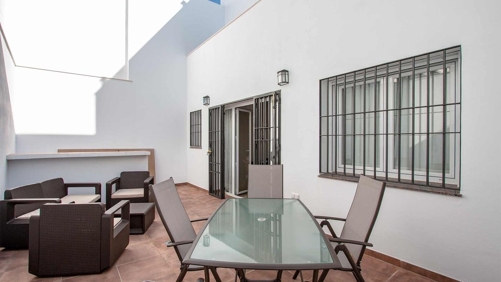 A modern outdoor patio with a glass table and chairs, white walls, and barred windows.