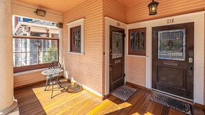 Une véranda couverte avec un plancher en bois, une table en métal et des chaises. On y trouve deux portes dotées de panneaux de verre et un numéro au-dessus de chacune.