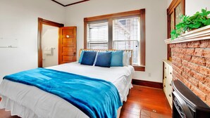Une chambre à coucher comprenant un lit, une commode, une cheminée en briques et une fenêtre avec des stores.