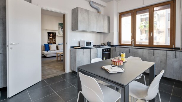 Cocina moderna con armarios grises, un microondas y una estufa. Hay una mesa de comedor con sillas y un ventanal con marcos de madera.
