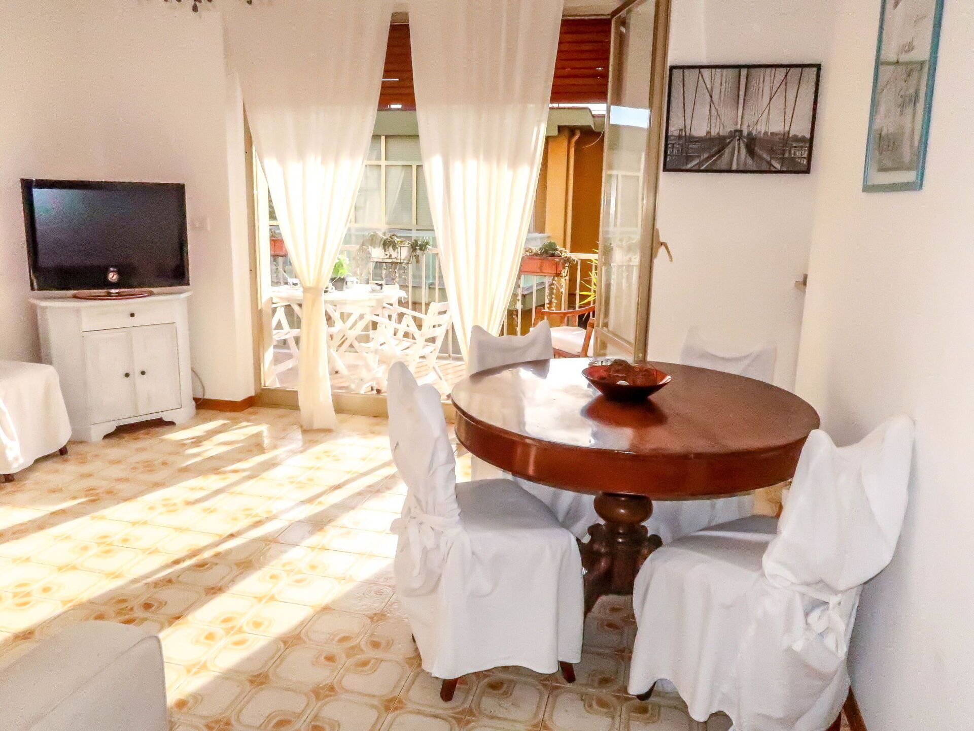 Un comedor con una mesa redonda, sillas blancas, un televisor sobre un mueble y vistas al exterior.