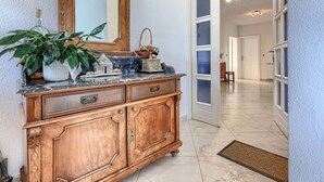 Um corredor com um armário de madeira, um espelho e uma planta. Uma porta dá para outro quarto.