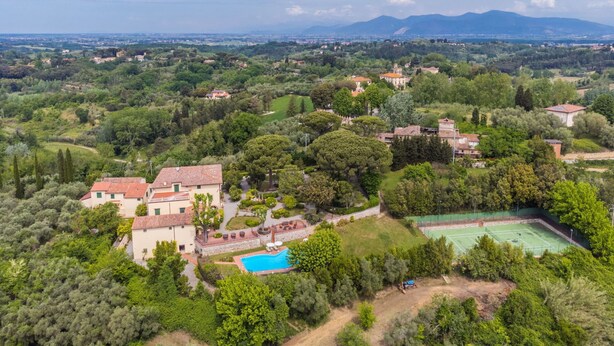 Una vista aerea di una villa con piscina, campo da tennis e rigogliosa vegetazione circostante.