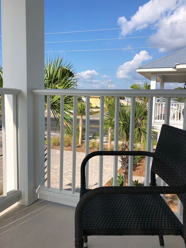 Un balcon avec un fauteuil en osier, des balustrades blanches et une vue sur des palmiers et un immeuble.