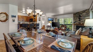 A dining area with a wooden table set for six, surrounded by chairs. A kitchen and living area are visible in the background. - Managed by Park City Lodging-True Ski-In/Ski-Out to Park City Mountain Resort! (Park City)