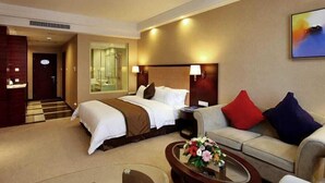 A hotel room with a bed, a sofa, a coffee table with flowers, a TV, and a bathroom visible through a glass door. - Jiayue Hotel (Shenzhen)