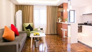A modern hotel room with a sofa, a small table with fruits, a chair, and a kitchenette. - Jiayue Hotel (Shenzhen)