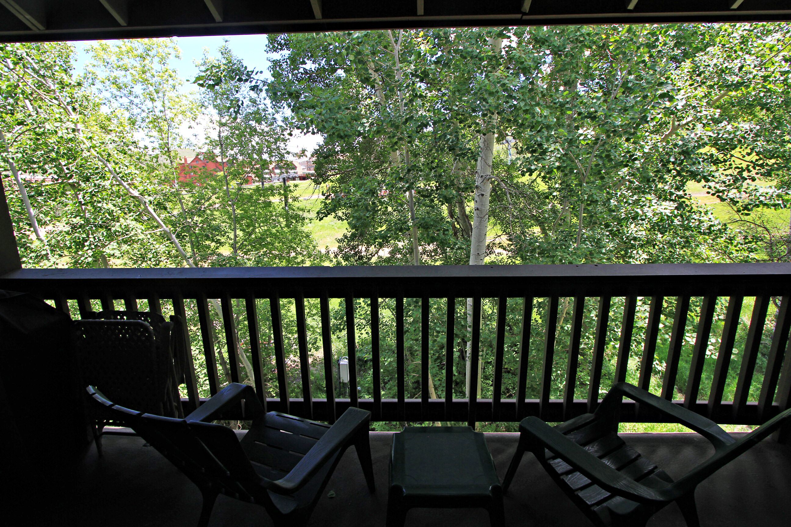 A balcony with two chairs and a small table, overlooking a green area with trees and a building in the background.