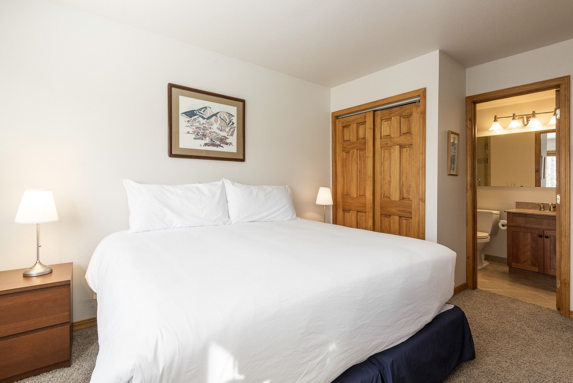 A neatly made bed with white linens, a wooden wardrobe, a framed picture on the wall, and a bathroom visible through an open door.