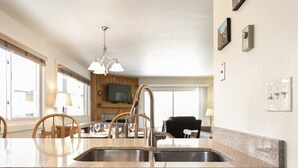 Una cocina con encimeras de granito, pileta de acero inoxidable y un grifo moderno. Se observa un comedor al fondo con una mesa y sillas de madera.