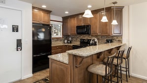 A modern kitchen with wooden cabinets, a black refrigerator, a microwave, a stove, and a granite countertop. There are three pendant lights and a bar stool with a cushioned seat. - Managed by Park City Lodging-True Ski-In/Ski-Out to Park City Mountain Resort! (Park City)