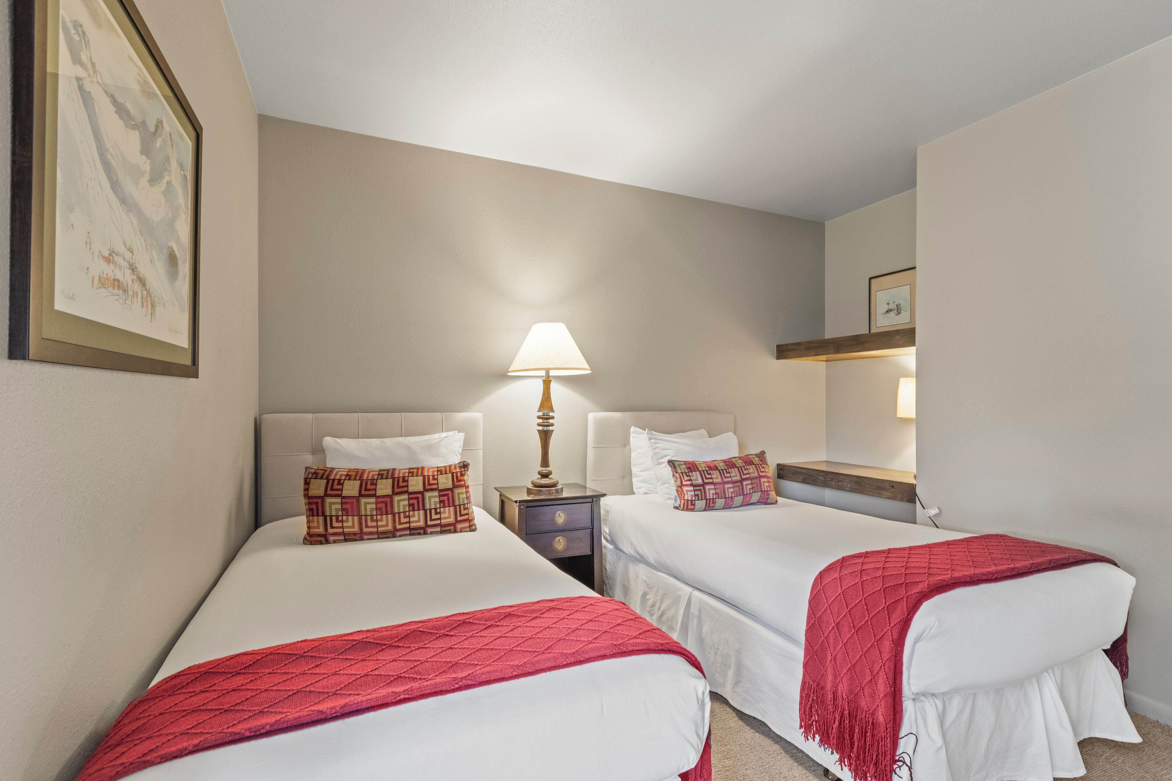 Una camera d'albergo con due letti, un comodino con una lampada, un quadro appeso alla parete e un piccolo ripiano con uno specchio.