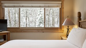 Ein Schlafzimmer mit einem Bett, einem Fernseher auf einem Holztisch, einer Lampe und einem Fenster mit Blick auf schneebedeckte Bäume.