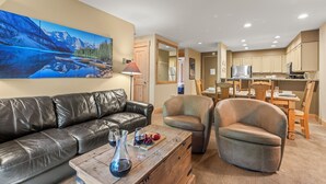 Ein Wohnzimmer mit einem schwarzen Ledersofa, zwei Ledersesseln, einem Couchtisch aus Holz mit Weingläsern und Früchten sowie einem Gemälde einer Berglandschaft.
