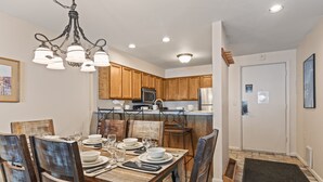 A dining area with a wooden table set for four, surrounded by wooden chairs. A chandelier hangs above, and the kitchen is visible in the background with wooden cabinets and modern appliances. - Managed by Park City Lodging-True Ski-In/Ski-Out to Park City Mountain Resort! (Park City)