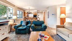 A spacious living room with two teal armchairs, a wooden coffee table with wine glasses, and a beige sofa. A kitchen with wooden cabinets and a dining area is visible in the background. - Managed by Park City Lodging-True Ski-In/Ski-Out to Park City Mountain Resort! (Park City)