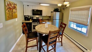 A kitchen with white cabinets, a microwave, stove, and refrigerator. There is a dining table with chairs and a window with blinds.