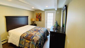 A bedroom with a large bed, a nightstand, a desk with a computer, a painting on the wall, and a door with a window.