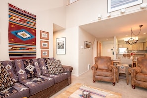 A living room with a sofa, armchairs, and a coffee table. There is a kitchen in the background.