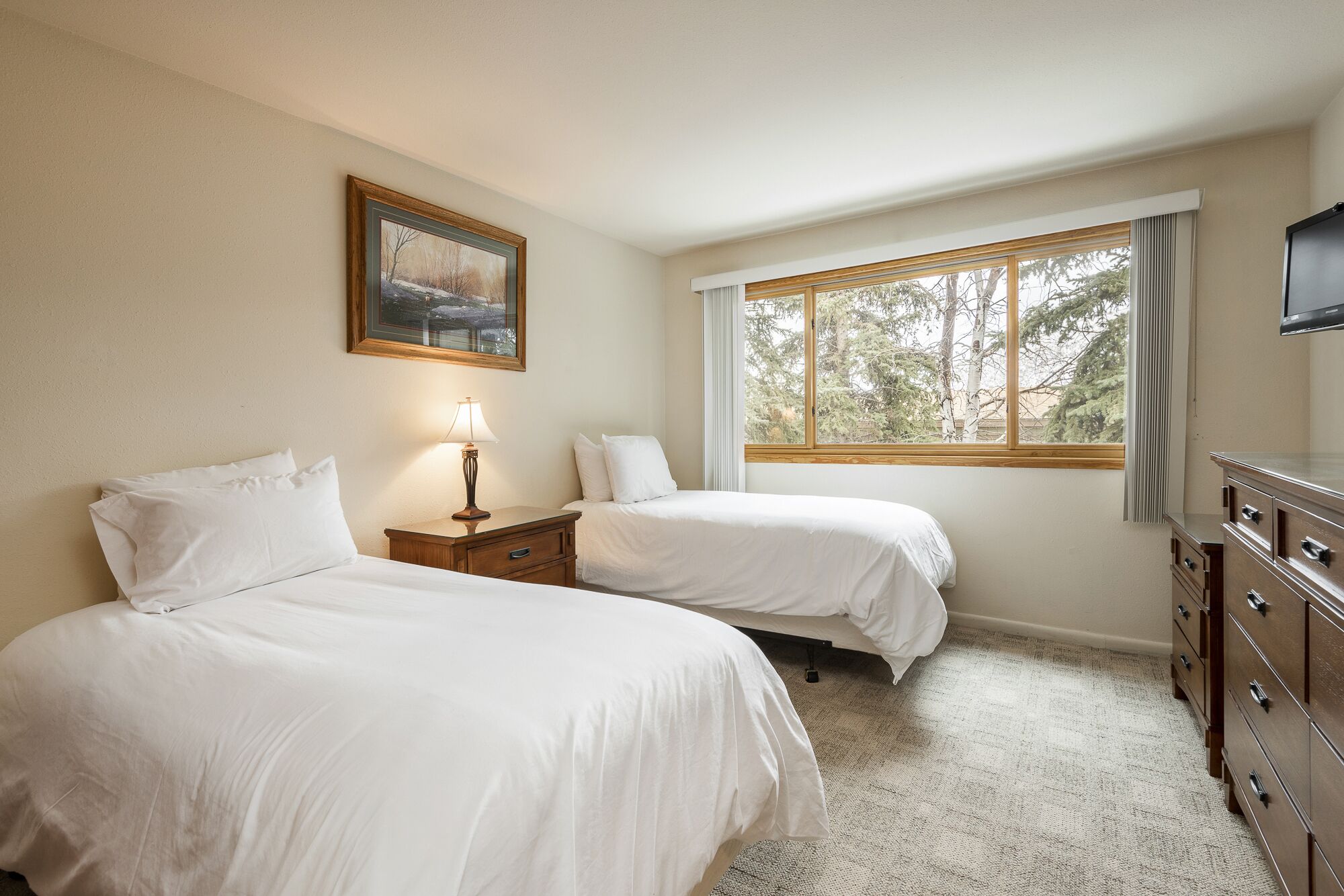 A hotel room with two beds, a nightstand with a lamp, a dresser, a painting on the wall, and a window with a view of trees.