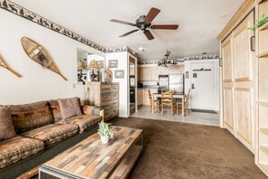 Uma sala de estar acolhedora com uma mesa de centro de madeira, um sofá com almofadas estampadas, e um espaço de jantar com cadeiras de madeira, com uma cozinha ao fundo.