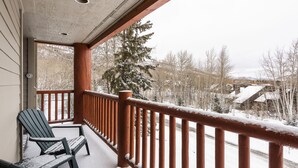 Ein überdachter Balkon mit Holzgeländern und zwei Adirondack-Stühlen, mit Blick auf eine verschneite Landschaft mit Bäumen und Häusern.