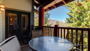 A covered outdoor area with a round glass table and chairs, wooden railings, and a view of trees and mountains.