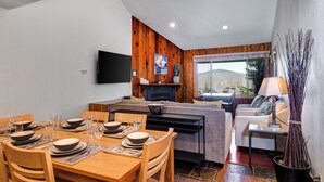A modern living room with a dining area, wooden panel walls, a fireplace, and a view of the outdoors.