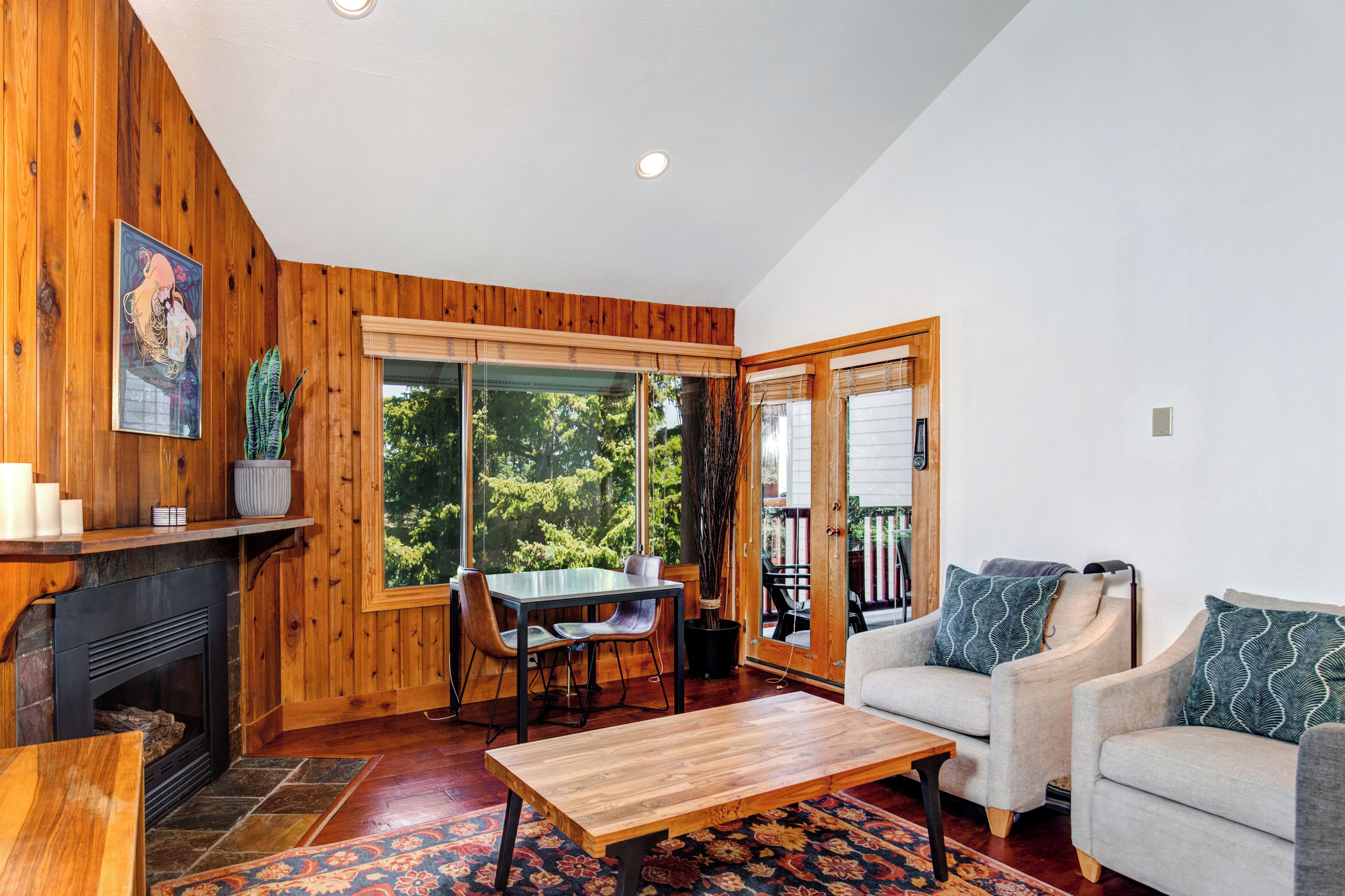 A cozy living room with wooden walls, a fireplace, a dining table with chairs, a sofa, and a large window offering a view of greenery.