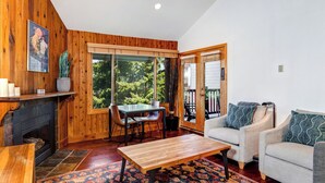 A cozy living room with wooden walls, a fireplace, a dining table with chairs, a sofa, and a large window offering a view of greenery.