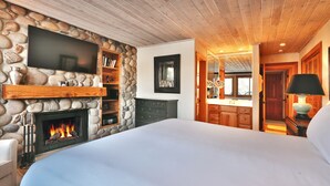 Un dormitorio con chimenea de piedra, televisor de pantalla plana, techo de madera y vista a la cocina.