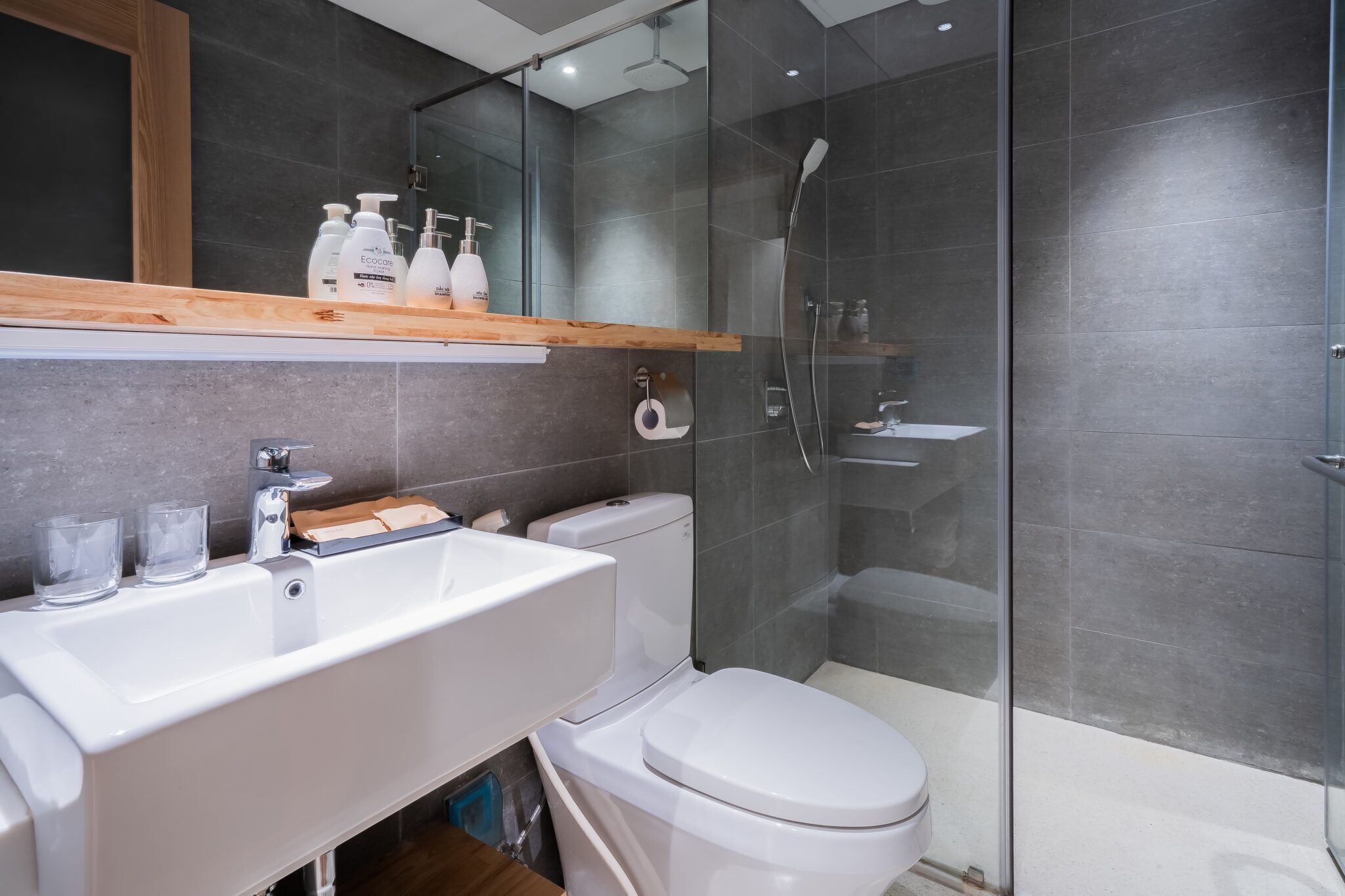 Une salle de bain moderne avec un lavabo, des toilettes et une douche, le tout de couleur blanche. On peut voir des bouteilles sur le lavabo et un miroir au-dessus.