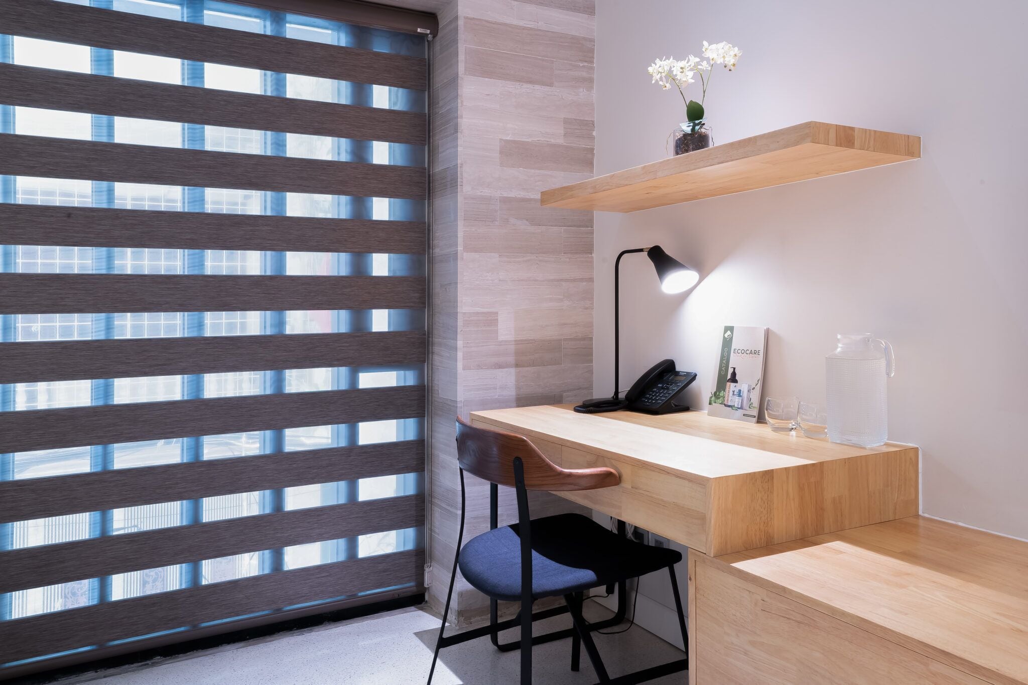 A modern office space with a wooden desk, a chair, a lamp, a phone, a vase with flowers, and a water pitcher.