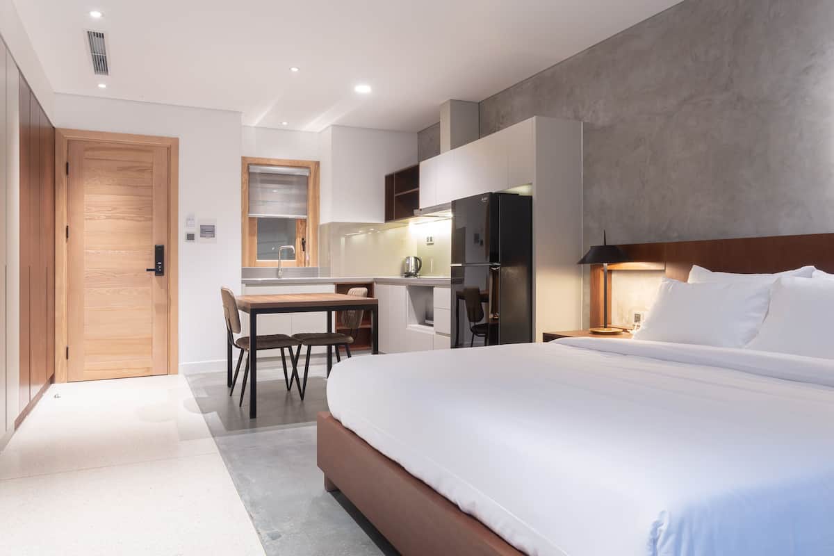 Een moderne hotelkamer met een groot bed, een bureau met stoel, een kitchenette en een houten deur.