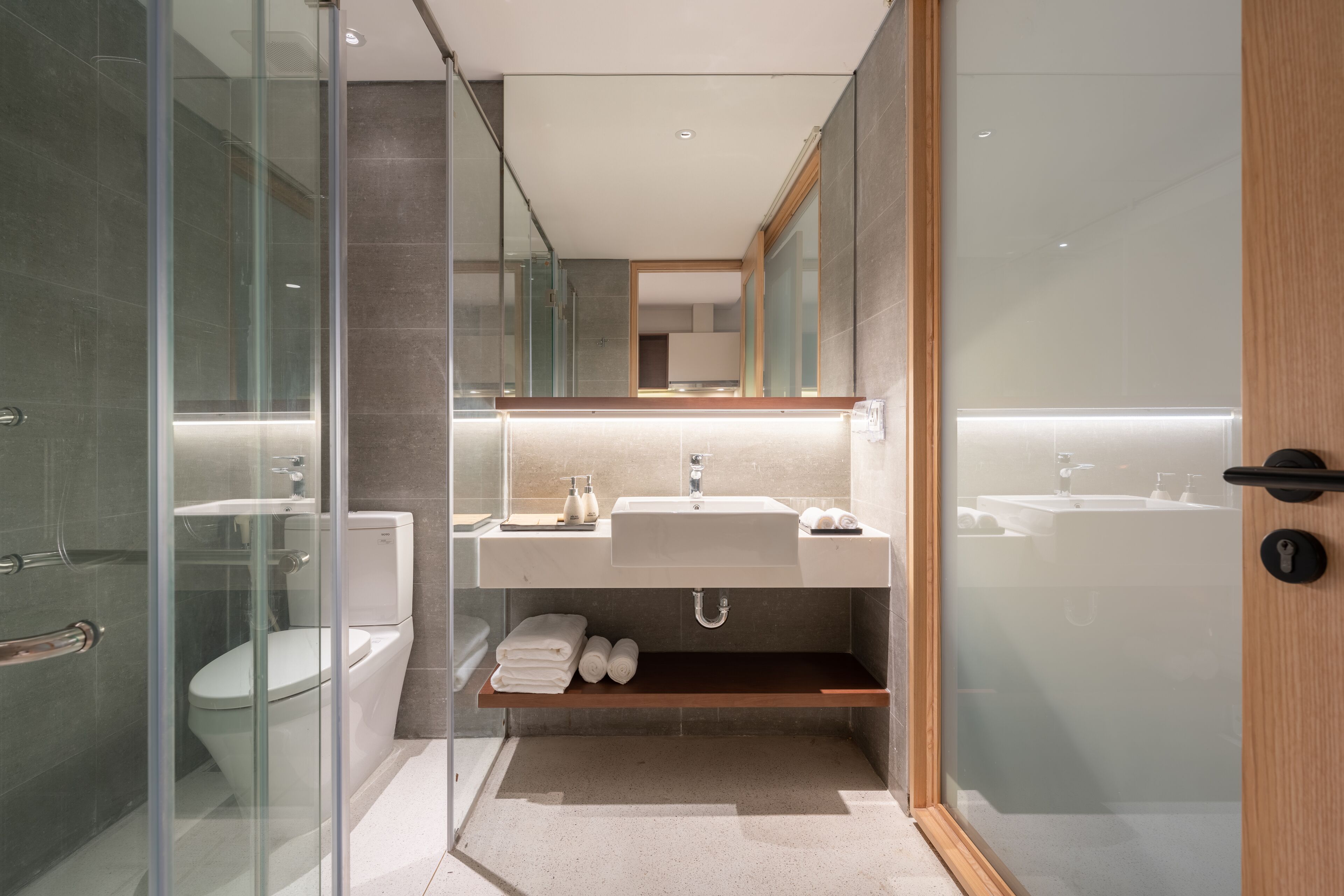 A modern bathroom with a glass shower enclosure, a white sink, and a wooden shelf with towels.