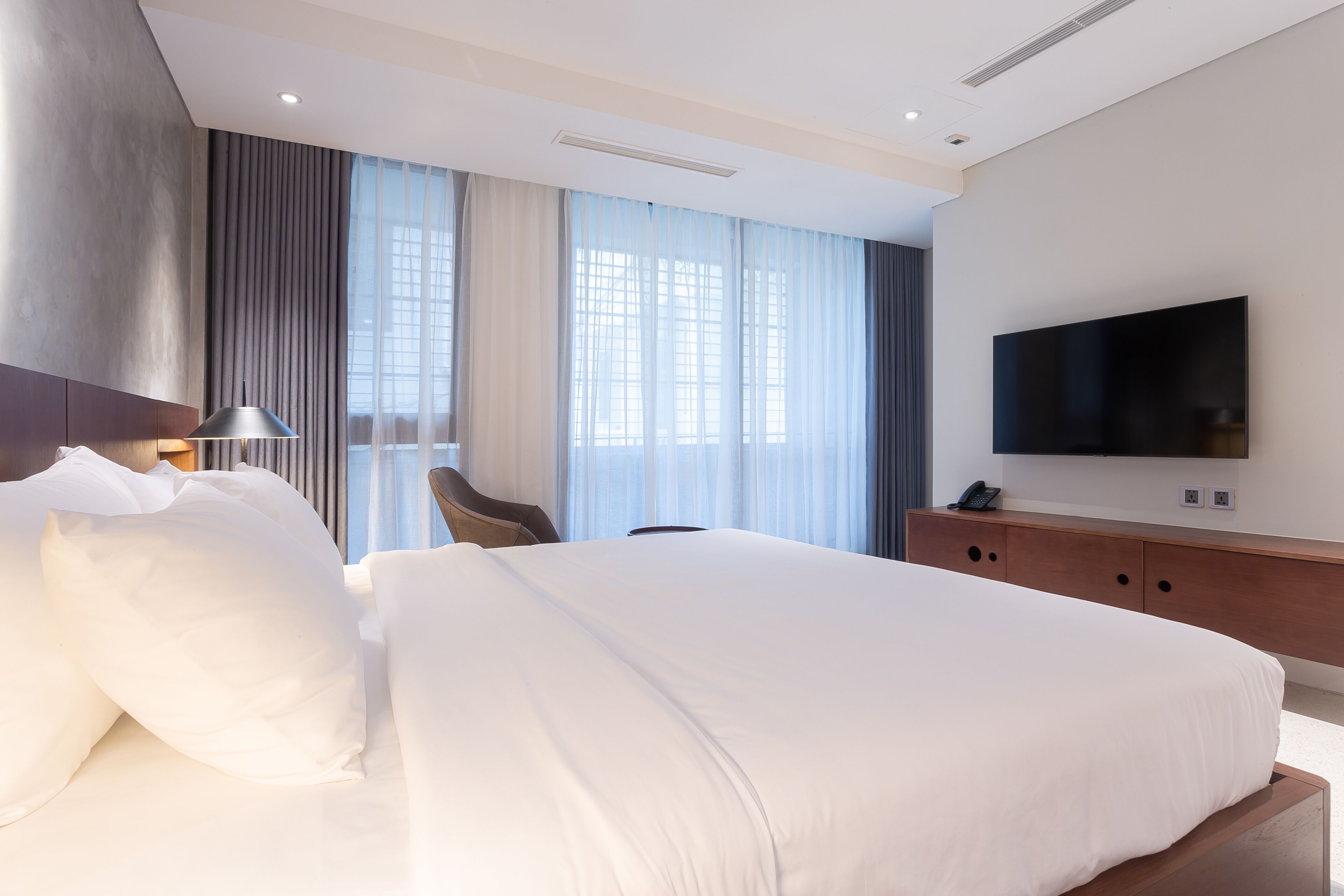 A modern hotel room with a large bed, a flat-screen TV, a wooden dresser, and a large window with curtains.