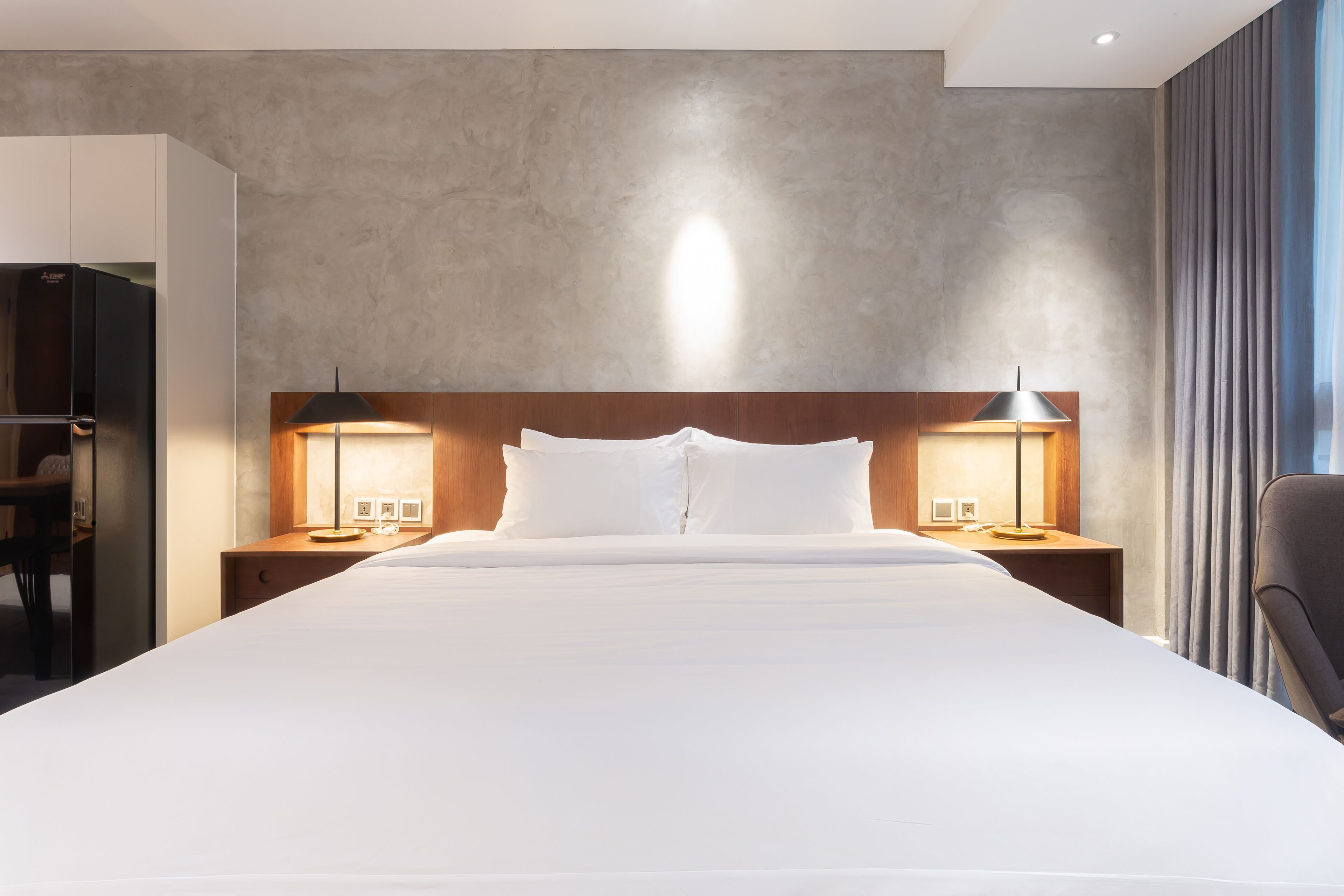 Ein modernes Hotelzimmer mit einem großen Bett, zwei Nachttischen inklusive Lampen, einem hölzernen Kopfteil und Blick auf die Stadt durch große Fenster.