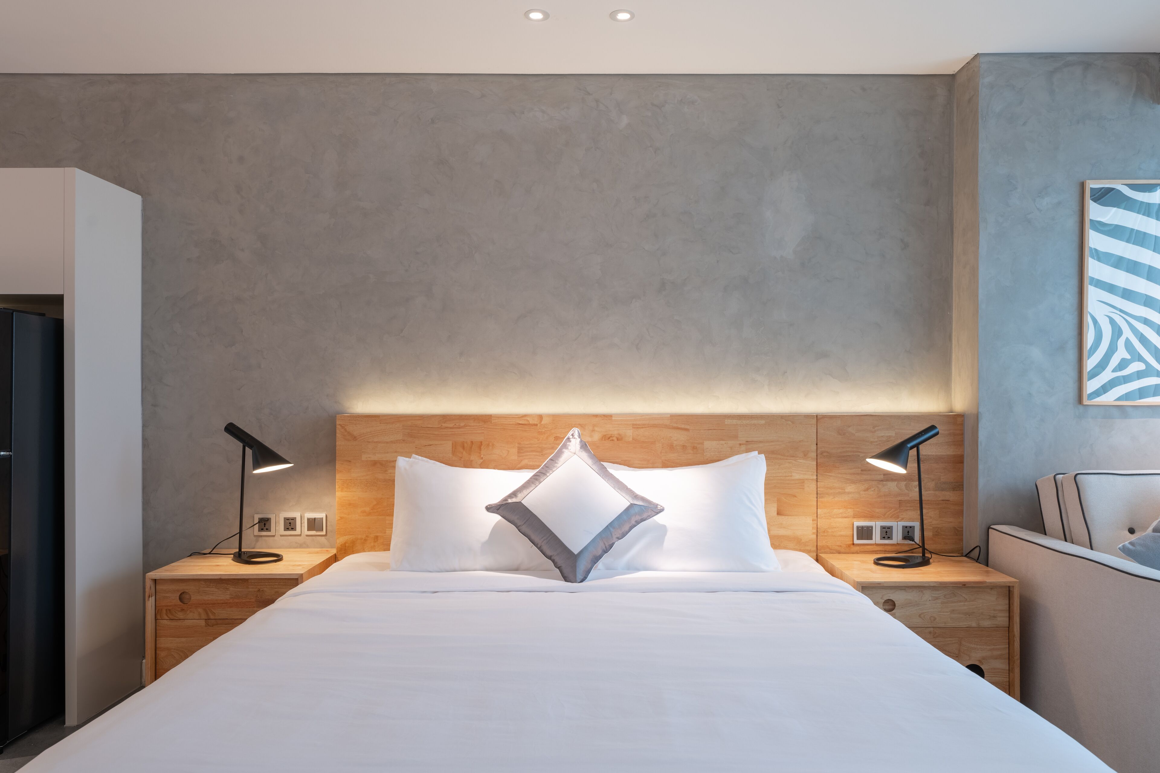 A modern hotel room with a large bed, wooden headboard, two bedside tables with lamps, and a grey textured wall.